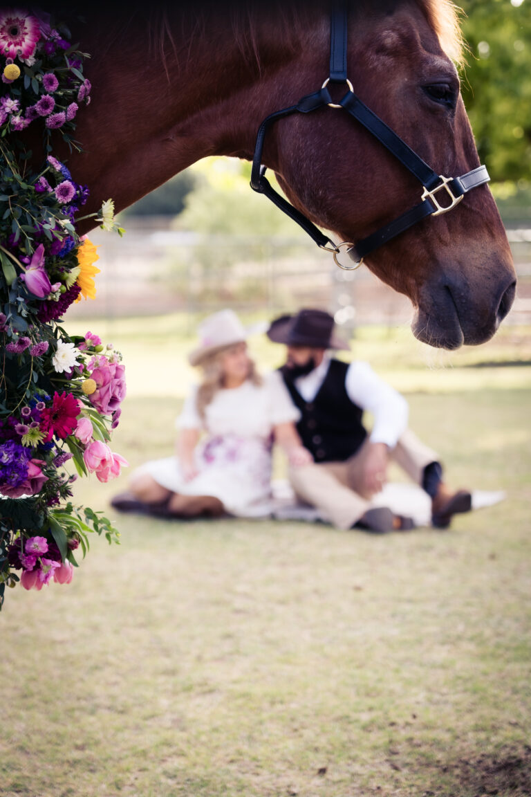 Couple in Surprise AZ at Freedom Farms blurry in the background wearing western clothing with the head of a horse.