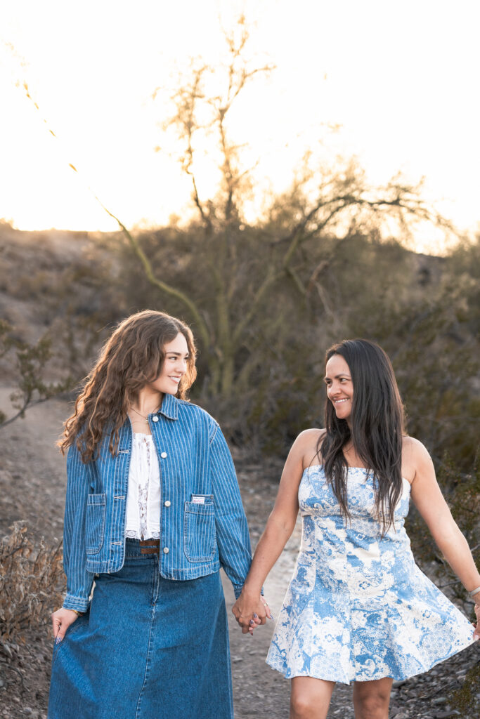 Graduate in a denim jacket and denim skirt holding hands with her mom in a blue and white dress walking in the Arizona desert at Lake Pleasant.